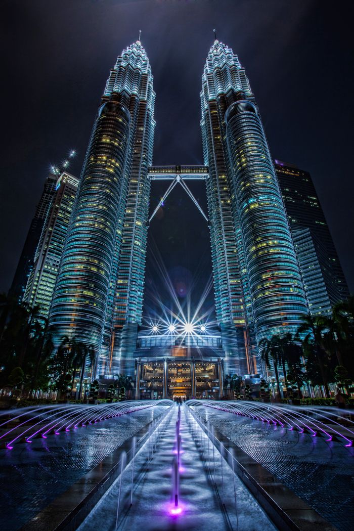 Dramatic night shot of Petronas Towers in Kuala Lumpur with illuminated fountains in foreground.